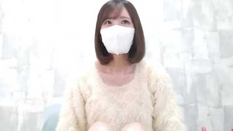 Video of an Asian woman with shoulder-length brown hair, wearing a white face mask and a fluffy, beige sweater, seated indoors with semi-transparent white curtains in the background.