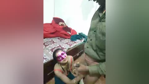 Video of a young boy with a Superman mask, wearing a red shirt, kneeling in a messy room with a red blanket, a green shirt, and a wooden bed.