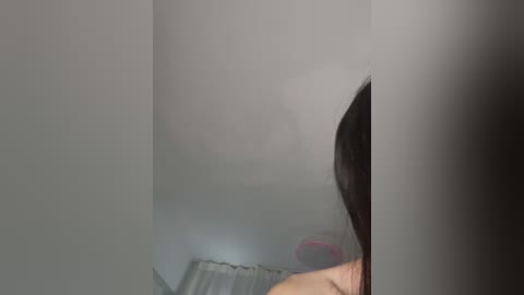Video of a person's upper body reflected in a mirror, showing a dark-haired individual's face and shoulders. The background includes a light-colored wall and a hint of a pink object, possibly a pillow, in the lower left corner.