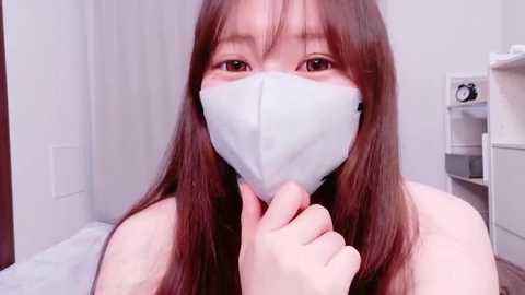 A video of an Asian woman with long, straight brown hair, wearing a white face mask, holding it with her left hand. She is indoors, with a white wall and a white shelf in the background.