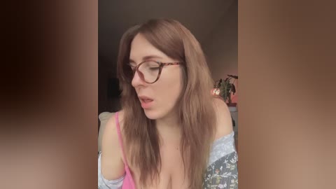 A video of a young woman with long, straight brown hair, fair skin, and wearing glasses, captured in a casual, off-shoulder floral top, indoors with blurred background.