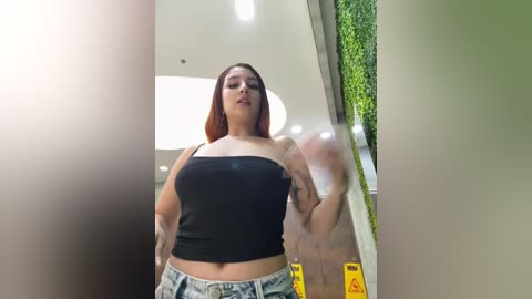 Video of a young woman with medium-length red hair, wearing a black crop top and light-wash jeans, captured indoors near a window with greenery outside.