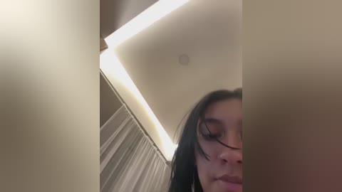 Video of a young woman with long, dark hair, partially obscured by a beige curtain. She has fair skin and a neutral expression. The background features a minimalist, modern interior with soft lighting and white walls.