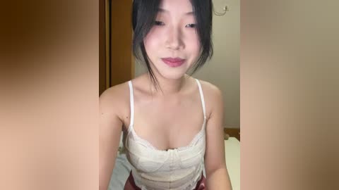 Video of an East Asian woman with light skin and straight black hair, wearing a white lace camisole, standing in a dimly lit room with wooden paneling and a beige wall.