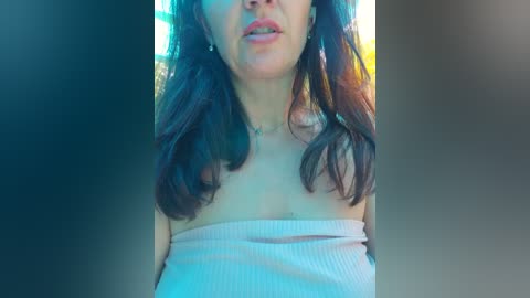 Video of a woman with medium skin tone and dark brown hair, wearing a strapless, ribbed top, looking down with slightly parted lips. Background is a blurred gradient of blue and green.
