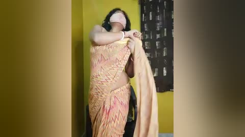 A video of a woman with long black hair, wearing a yellow and pink sari, partially covering her face, standing against a yellow wall with a black window shade.