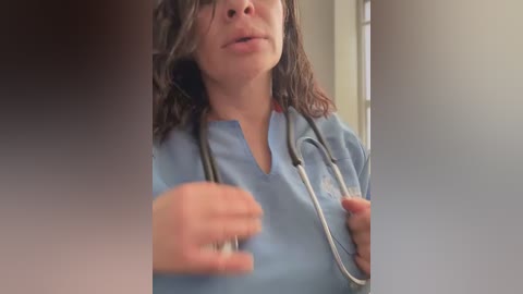 A video of a woman in blue scrubs, with shoulder-length brown hair, speaking with an open mouth. She holds a stethoscope around her neck, and a blurred figure is partially visible in the foreground.