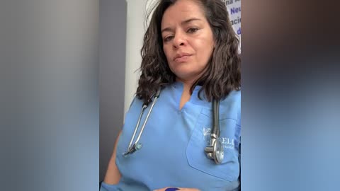 A video of a middle-aged woman with shoulder-length dark hair, wearing a blue medical scrub top, a stethoscope around her neck, and a serious expression. She stands in a hospital corridor, with a blurred background.