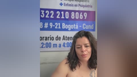 Video of a woman with shoulder-length dark hair, fair skin, and tattoos, standing in front of a hospital room wall with phone numbers and hospital information.