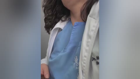 Video of a woman with wavy brown hair, wearing a white lab coat over a light blue scrub top, partially visible face, blurred background.
