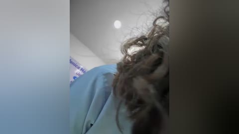 A video captures a person with curly brown hair peering through a doorway. The background shows a blue wall and part of a white ceiling with a light bulb. The setting appears clinical, possibly a hospital.