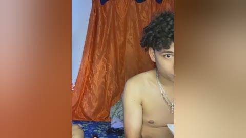 Video of a young man with curly hair and light brown skin, topless, standing in front of an orange curtain and blue fabric backdrop, wearing a silver chain necklace.