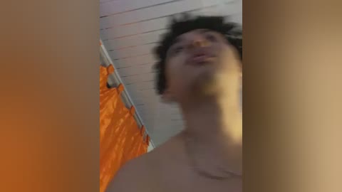 A blurred video of a shirtless person with dark hair, standing in a room with an orange curtain and white ceiling. The image is out of focus, making it difficult to discern details.