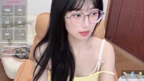 Media: A video of a young Asian woman with long black hair, wearing glasses and a yellow off-shoulder top, sitting on a chair.