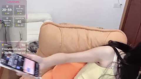 Video of a woman with fair skin and long black hair, wearing a sleeveless yellow top, reclining on a beige cushion. She's holding a smartphone, capturing a selfie. Background shows a white couch and wooden door.