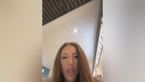 A video of a young woman with long, straight brown hair and fair skin, wearing minimal makeup, taken indoors near a large, reflective surface that distorts her face.