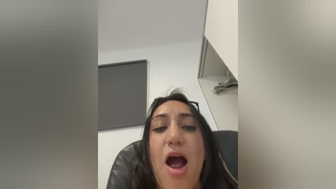 Video of a woman with long black hair, wearing black glasses, sitting on a black chair in a white room. She has a surprised expression, mouth open, with a black wall and window in the background.