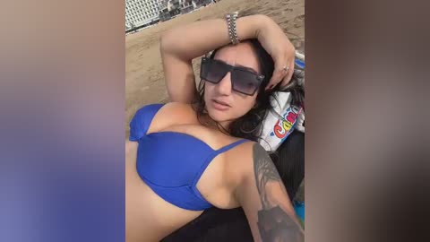 Video of a woman with medium skin tone, long dark hair, wearing a blue bikini top, large sunglasses, and a bracelet, lying on a beach with a towel and sand in the background.