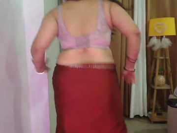 Video of a curvy, light-skinned woman with medium-length brown hair, wearing a lavender bra and a red skirt. She stands indoors, facing away, in a casual, slightly blurred setting with a wooden table, lamp, and curtains in the background.