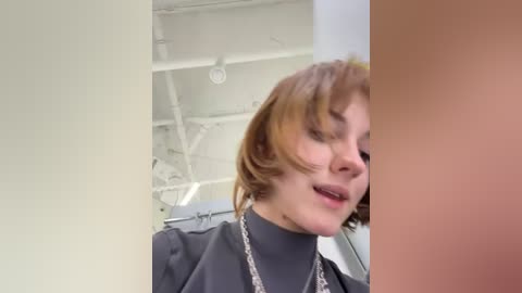 Media: Video of a light-skinned woman with short, reddish-brown hair, wearing a black shirt and silver necklace, standing in a white-tiled room with exposed pipes and a medical device in the background.