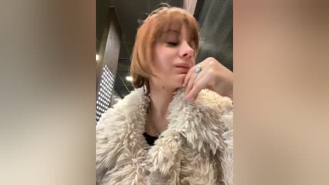 Video of a fair-skinned woman with short, reddish-brown hair, wearing a white, fluffy coat, indoors near a window with a grid pattern. She has a neutral expression and is slightly leaning forward.