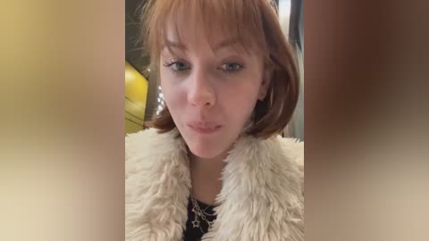 A close-up video of a young woman with fair skin, auburn hair, and a slight pout. She wears a fluffy white coat and a black star-patterned top. Background includes a blurred yellow object.