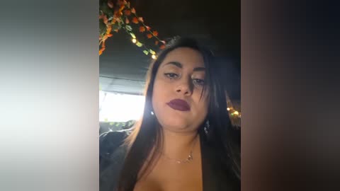Video of a young woman with long black hair, fair skin, and dark lipstick, wearing a black jacket, against a dimly lit background with a garland of orange leaves.