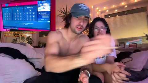 Video of a shirtless man and a woman with glasses, both smiling, sitting on a couch. TV screen in background shows a website. Warm, cozy room with string lights and plants.