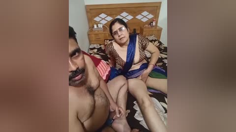 Media: Video of an Indian couple in a bedroom. The man, shirtless with a muscular build and dark hair, sits on a bed with a woman in a blue saree and glasses. Background features a wooden headboard and floral bedding.