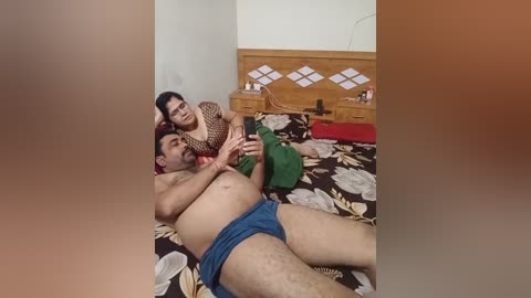 Video of a man lying on a floral bedspread, shirtless, wearing blue shorts, and a woman in a polka-dot top, sitting beside him, in a simple bedroom with wooden furniture.