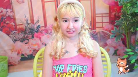 Video of a young, light-skinned, blonde-haired girl with curly hair, wearing a pink tank top with \"Air Fries\" text, sitting on a yellow chair against a floral wallpaper background.