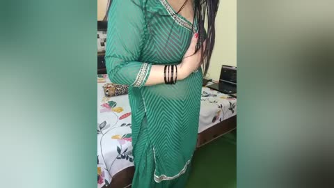 Video of a woman in a green, patterned, long-sleeved kurta, with black bracelets on her wrists, standing in a bedroom with a floral-patterned bedspread and green carpet.