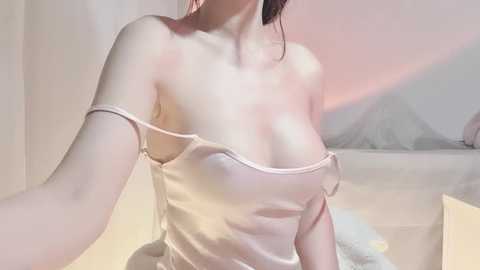 Video of a fair-skinned, slim woman with medium-sized breasts wearing a light pink, strapless satin camisole. Background features a soft, pastel-colored wall and a white, fluffy towel.