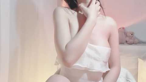 A soft, pastel-colored video of an Asian woman with fair skin, wearing a white, ruffled crop top, sitting on a bed with a plush teddy bear.