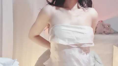 A video of a fair-skinned woman with long, dark hair, wearing a white towel that reveals her small breasts and toned abdomen, standing in a softly lit bedroom with a plush toy on a shelf.