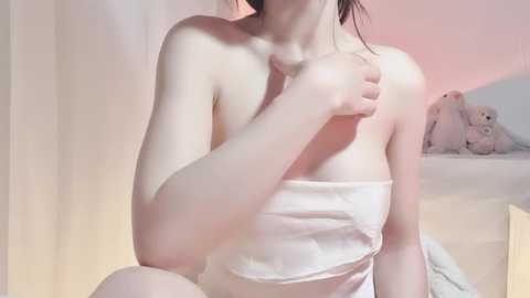 Video of a fair-skinned woman with dark hair, partially nude, holding a towel to her chest. Background features a teddy bear and soft, pastel-colored curtains.