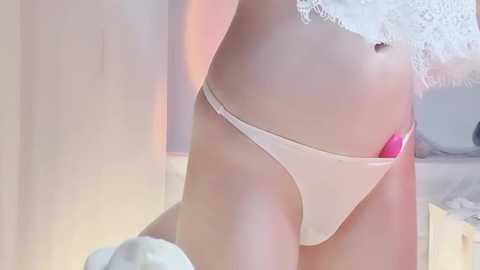 A close-up video of a woman in white lace lingerie, with a pink vibrator partially visible between her thighs, set in a softly lit bedroom with white curtains and a bed.