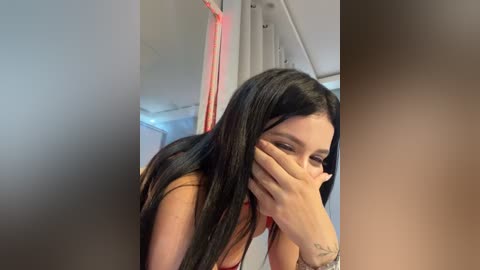 Video of a young woman with long black hair, wearing a tan top, covering her eyes, in a modern room with grey walls and a staircase.