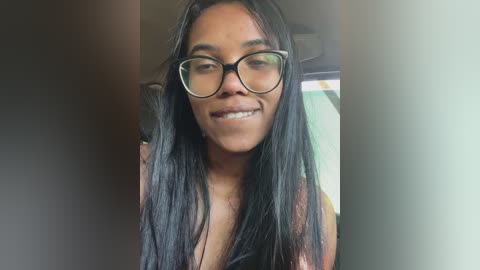 Video of a young Black woman with long, straight black hair, wearing black-rimmed glasses, and a black top, smiling. She stands indoors against a blurred background.