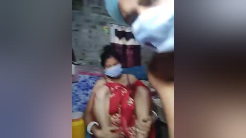 A blurry video depicts a woman in a red floral dress, seated on a blue and white patterned mat, with a man's legs around her, in a cluttered room with a concrete wall background.