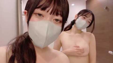 A video of an East Asian woman with long black hair, wearing a white face mask, standing topless in a bathroom, covering her breasts with one hand.