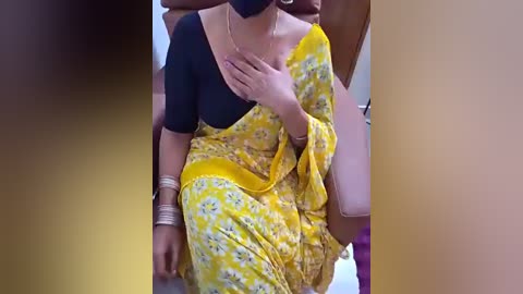 Video of a woman wearing a yellow sari with a black blouse, gold jewelry, and a black mask, sitting on a beige couch.
