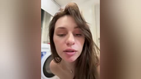 A video of a young woman with light skin and long brown hair, captured in a casual, slightly blurry bathroom setting. She wears a nose ring and has a neutral expression.