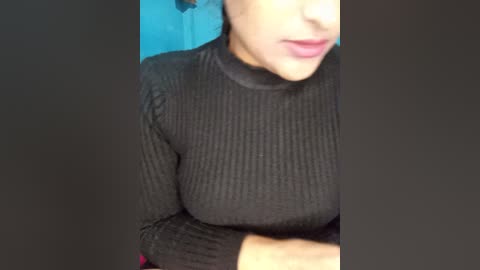 A close-up video of a woman wearing a black ribbed long-sleeve top, revealing her ample breasts. Her lips are slightly parted, and her skin tone is light. The background is blurred, with a teal wall.