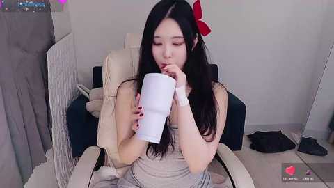 Video of an East Asian woman with long black hair and a red bow, wearing a grey dress, sitting on a white chair, holding a white mug, in a minimalist room with a blue sofa and a mirror.