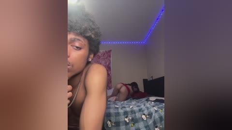 A video of a nude, dark-skinned person with curly hair, partially obscuring the view of a woman in red lingerie on a bed with blue and white checkered sheets, under blue LED lights.