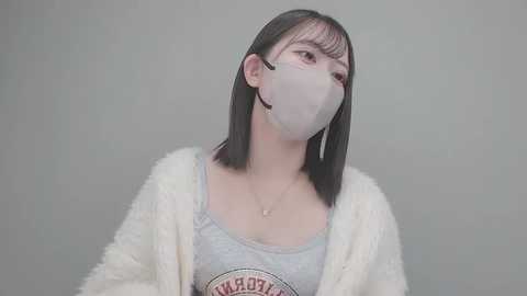Video of a young Asian woman with straight black hair, wearing a white fluffy cardigan over a grey tank top with a \"LACOSTE\" logo, and a white mask, against a plain light grey background.