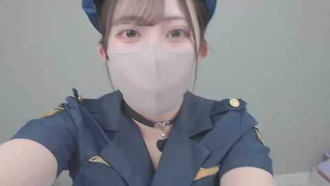Video of an Asian woman in a police uniform, wearing a white surgical mask, blue hat, and a black choker, with light skin and brown hair. Background is a plain, light-colored wall.