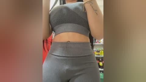 Video of a woman wearing a grey sports bra and high-waisted leggings in a supermarket aisle. Her left arm shows a tattoo. Shelves of products and a red shopping cart are in the background.