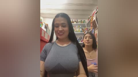 Video of two young women in a cluttered kitchen. One woman, with long dark hair, wears a tight grey top revealing ample cleavage. Another woman, with straight brown hair, stands in the background, looking towards the camera.